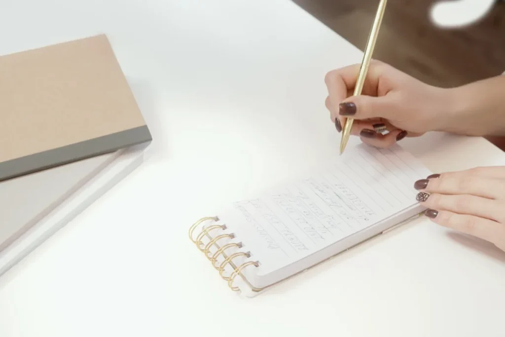 Hand schreibt mit goldfarbenem Stift in einen kleinen Spiralblock auf weißem Tisch