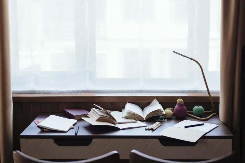 Schmaler Schreibtisch am Fenster mit aufgeschlagenen Büchern und Notizheften