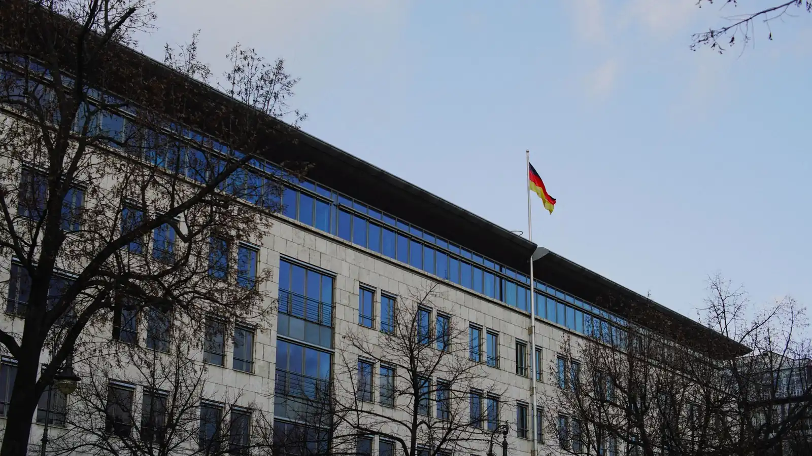 Bürogebäude einer Verwaltung mit deutscher Flagge, davor kahle Bäume unter blauem Himmel