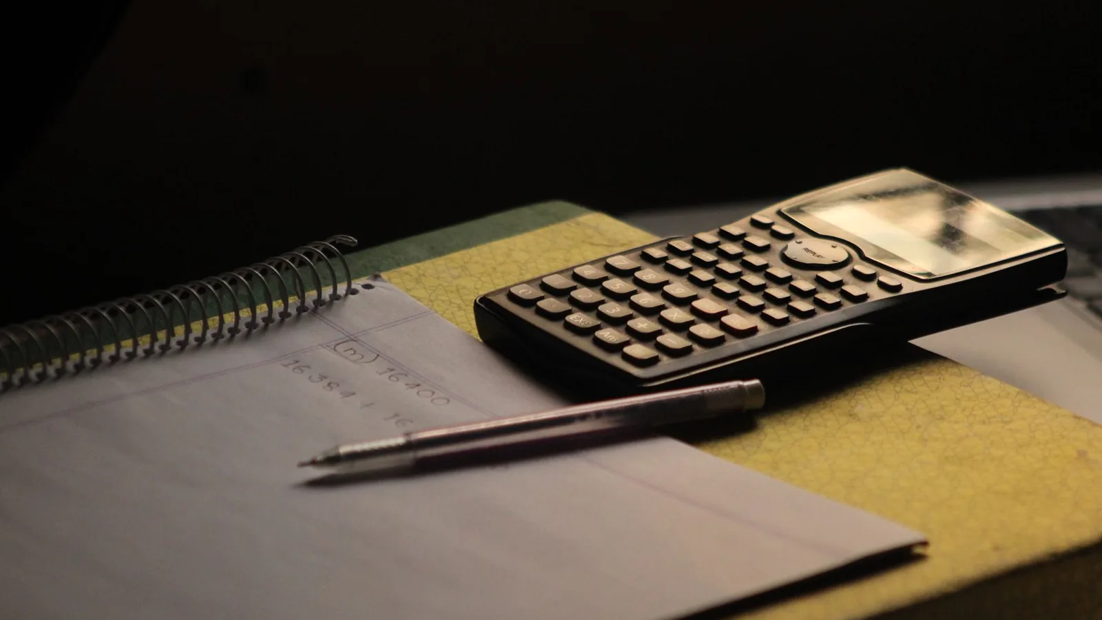 Taschenrechner, Stift und aufgeschlagenes Notizheft auf einem dunklen Tisch